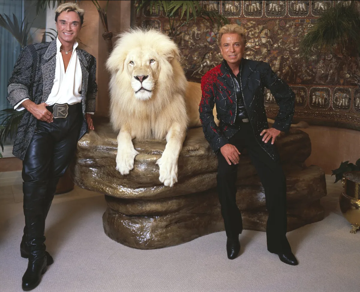Two men in black stage costume stand either side of a white lion lying on a rock.