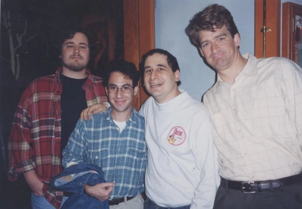 Four men stand close together in a room, smiling at the camera, in shirts and a sweatshirt.