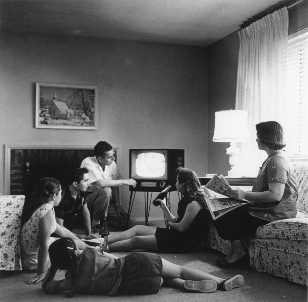 A father, mother and four children in a living room around a small television on thin legs; the mother holds a newspaper.