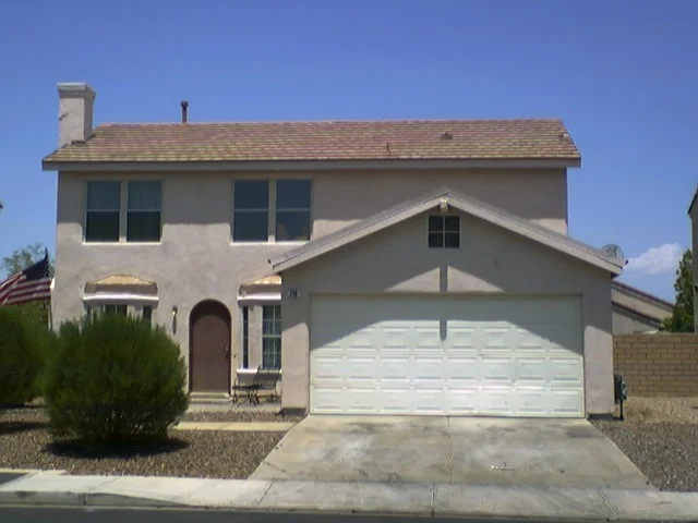 A plain two-storey beige house with a double garage and a low bush, under a blue sky.