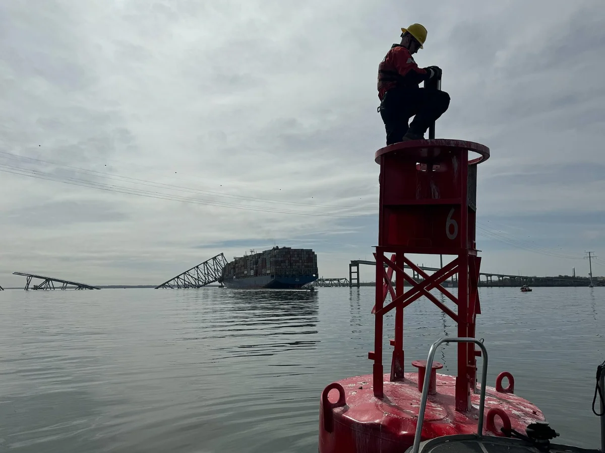 A worker stands on a red channel marker in grey water, with the fallen spans of the bridge and a container ship on the horizon behind.