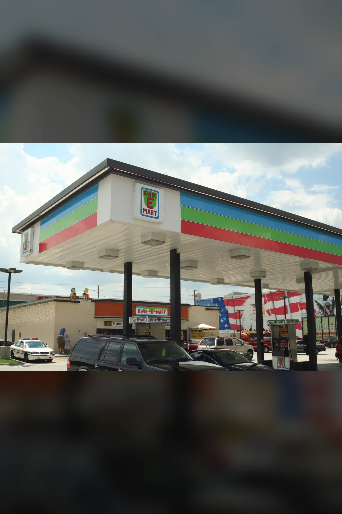 A filling station under a canopy striped blue, green and red with a Kwik-E-Mart sign. Cars stand at the pumps, and two small cartoon figures sit on the roof of the store behind.