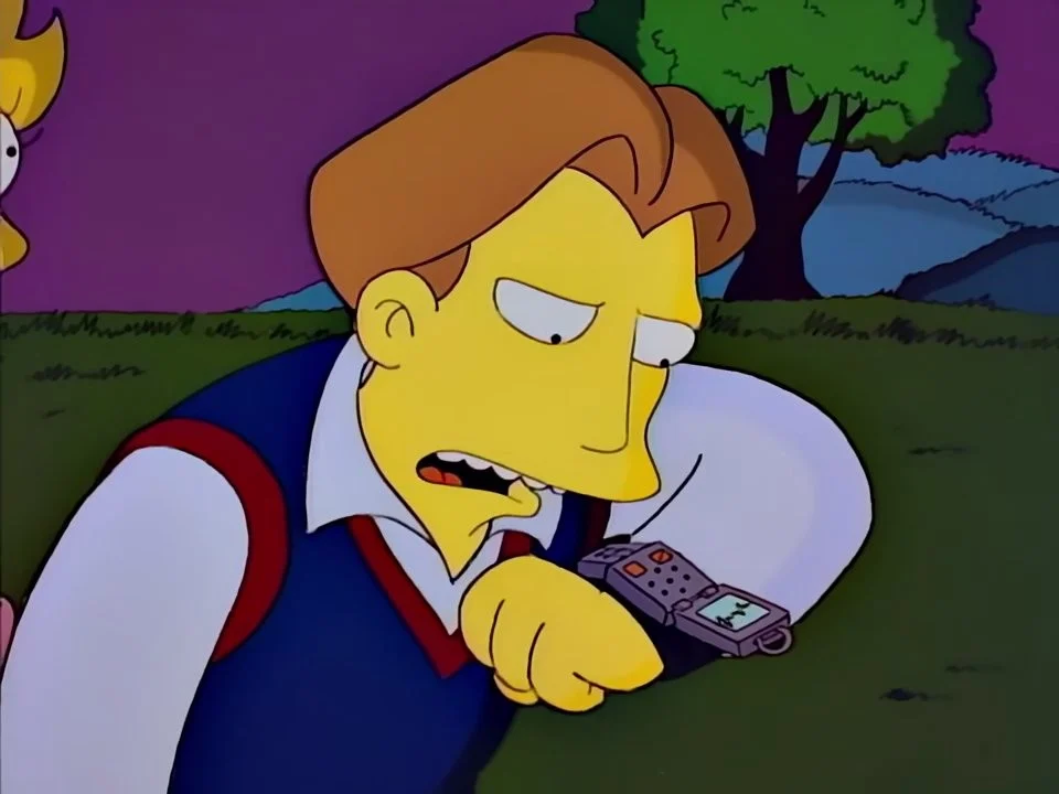 Hugh in a blue sweater vest bending his head to a chunky device strapped to his wrist.