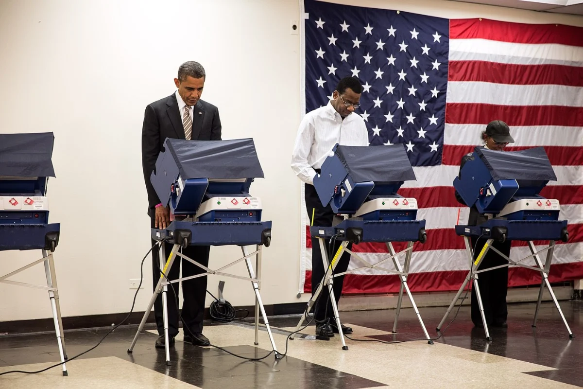 Barack Obama voting at an electronic machine on a stand, other voters beside him.