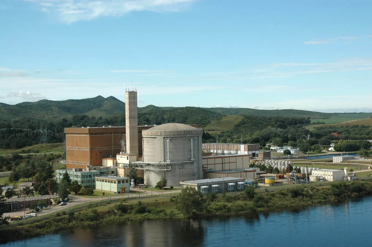 A reactor dome and stack beside a reservoir, hills behind.