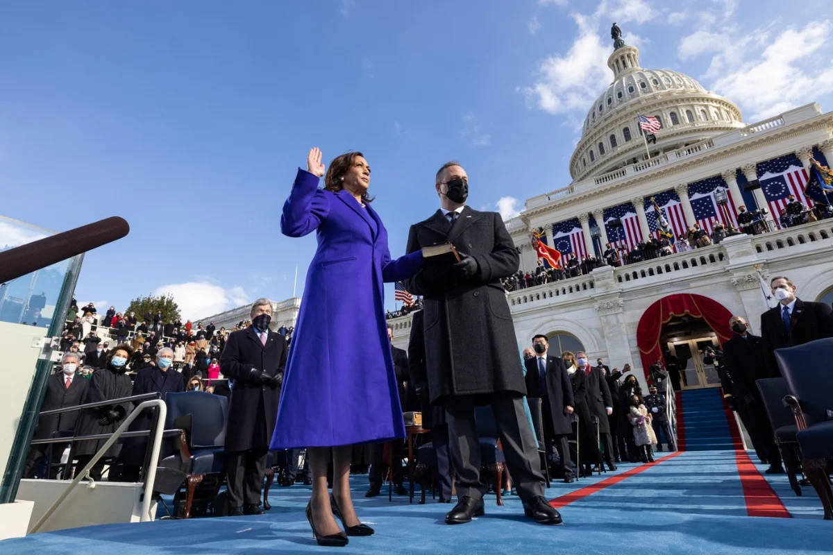 Kamala Harris in a purple coat with her right hand raised, the Capitol dome behind.