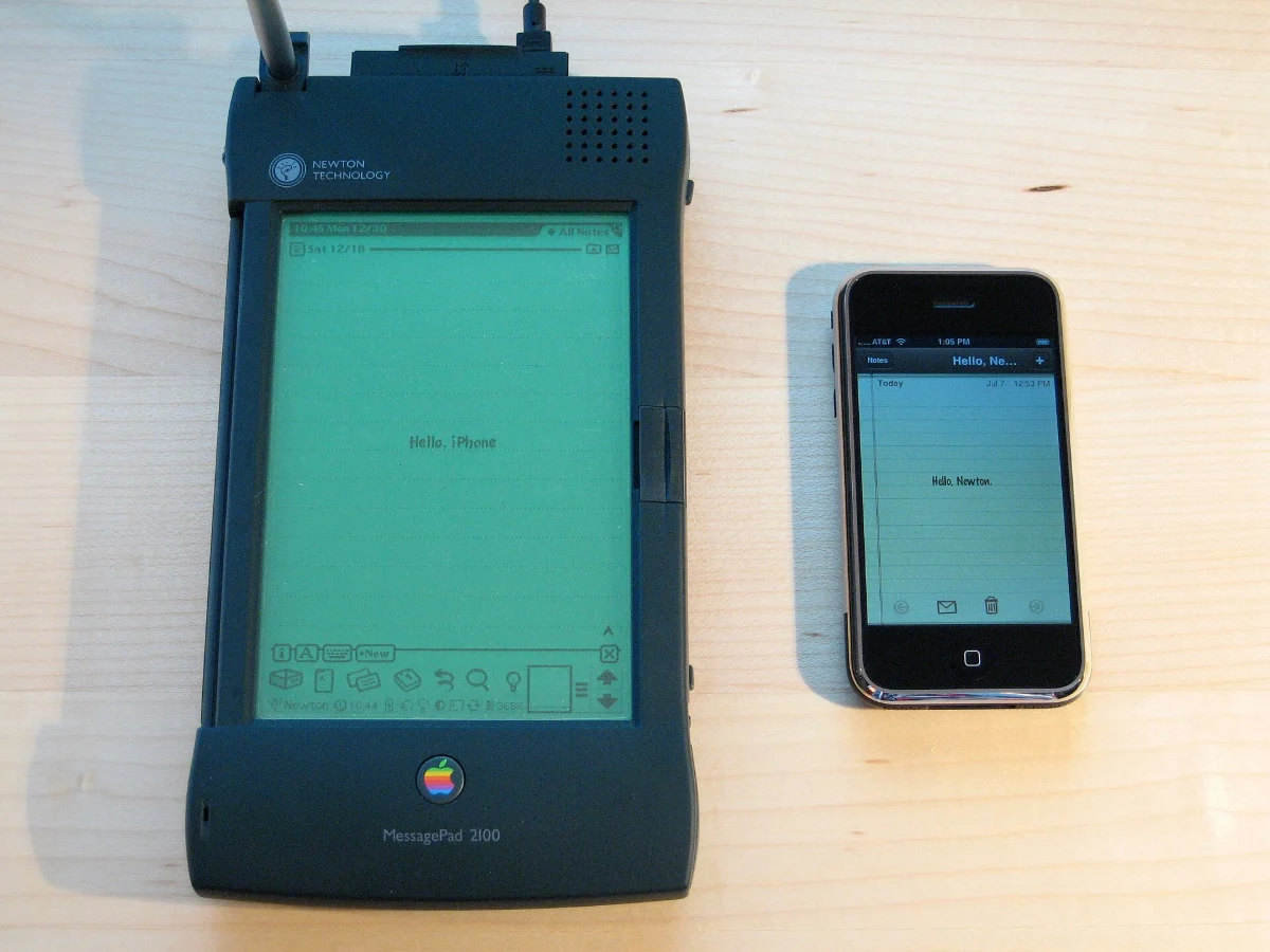 A large dark Apple Newton beside a much smaller iPhone, both lit, on a pale wooden table.