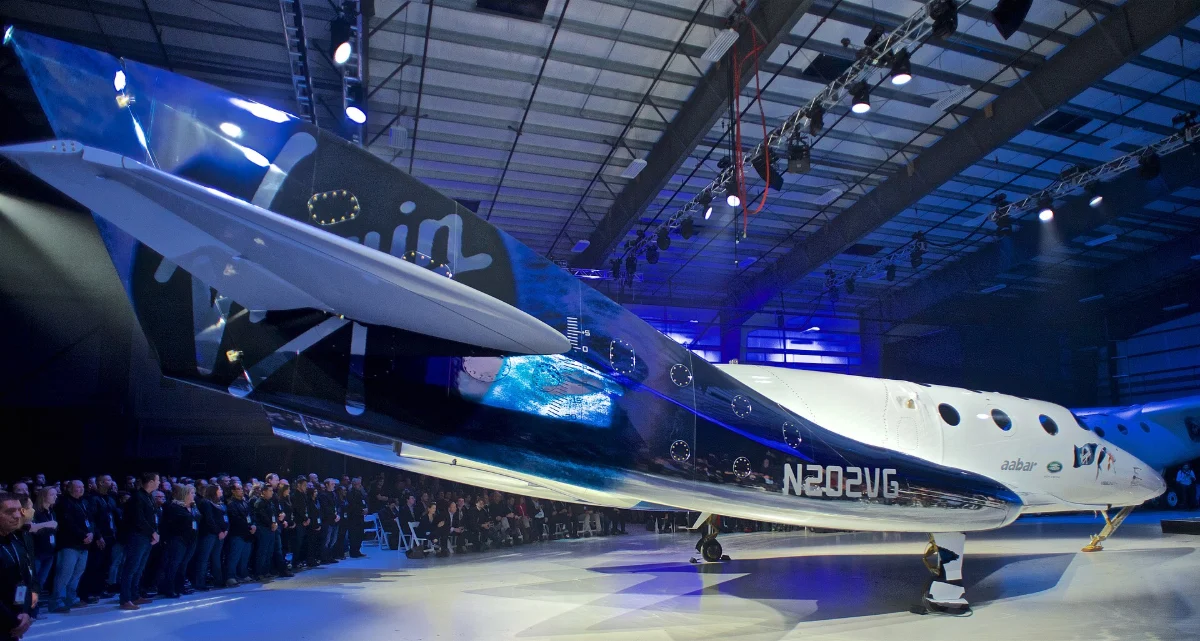 A white spaceplane under its carrier aircraft in a hangar lit blue, a crowd around it.