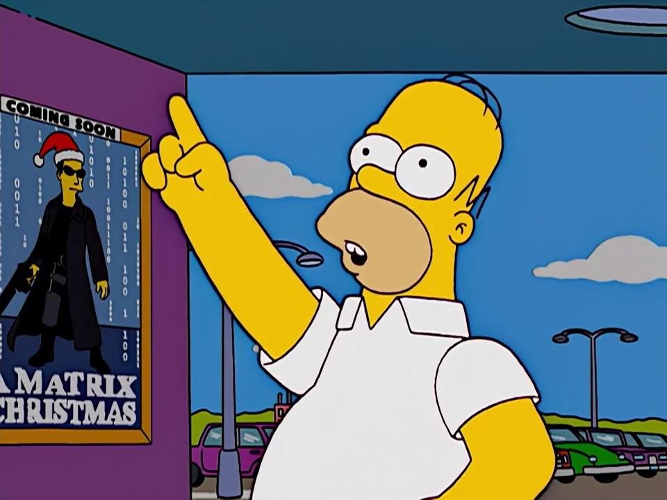 Homer at a ticket window pointing up, beside a framed poster of a figure in sunglasses, a long black coat and a Santa hat.