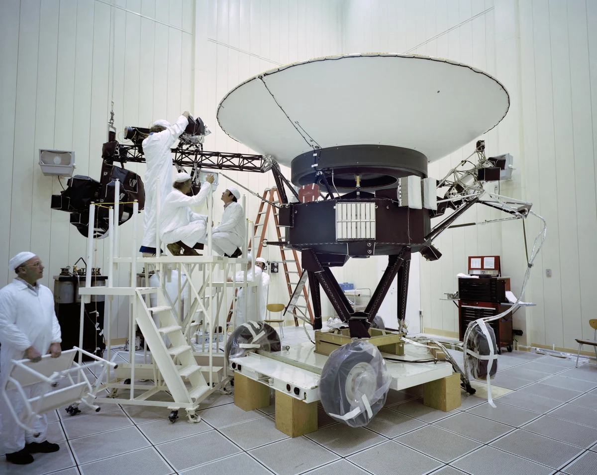 Technicians in white clean-room suits on platforms around a spacecraft with a large white dish antenna, in a white test hall