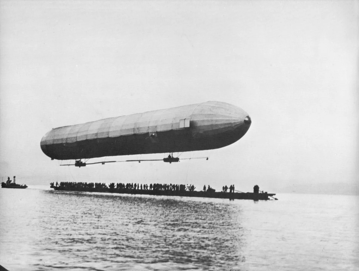 A long rigid airship with two small gondolas floating low above calm water, a line of dark figures on a raft underneath.