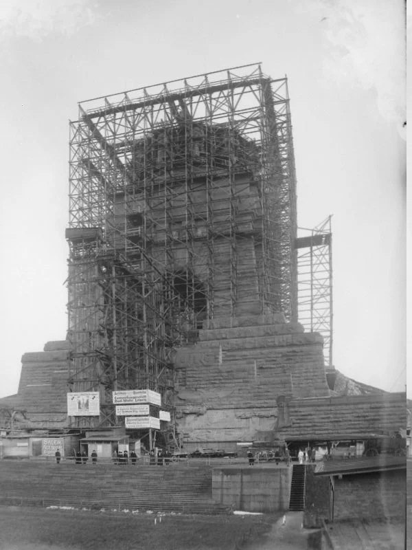 A stone tower wrapped from base to top in a tall timber scaffold, with advertising boards and small figures at its foot.