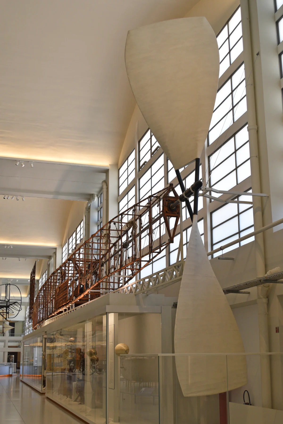 A very long narrow gondola framework hung high along the windows of a museum hall, with pale fabric blades at its end.
