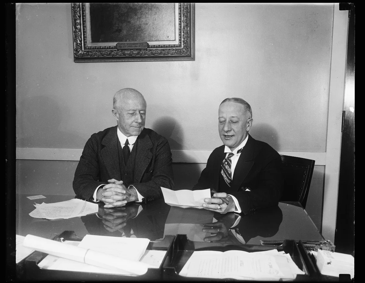 Two older men in dark suits seated side by side at a polished desk scattered with papers.
