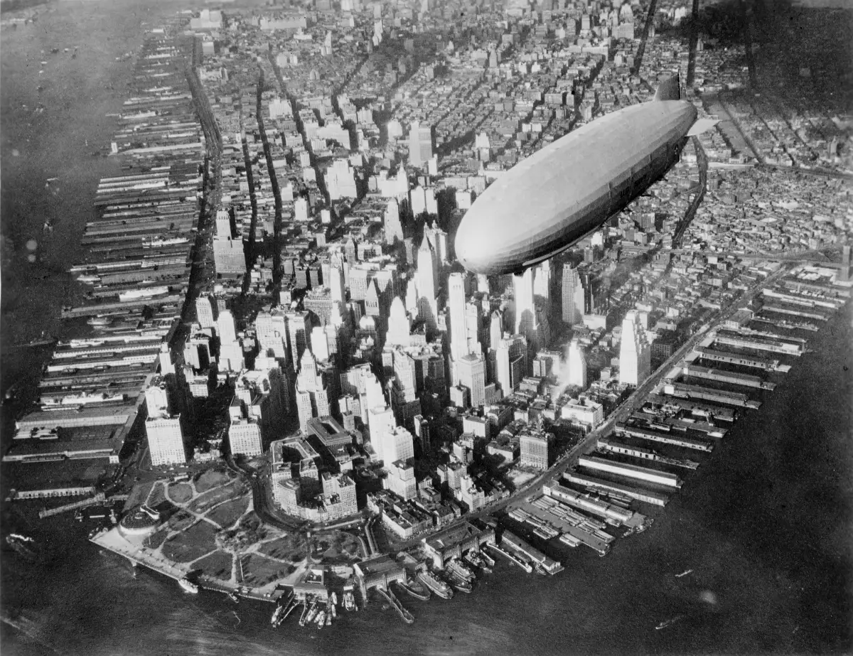 A huge silver rigid airship seen from above, flying over the southern end of Manhattan, with piers and rivers below.