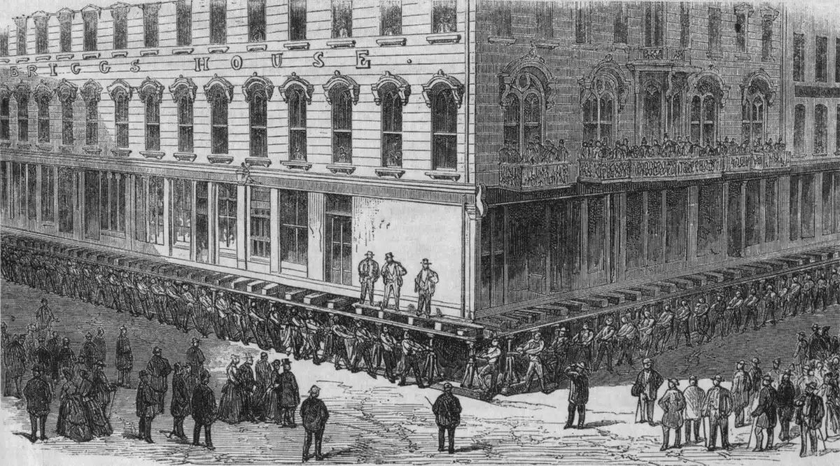 An engraving of a five-storey hotel on a street corner, raised on timbers, with dozens of men turning screws under it while a crowd watches.