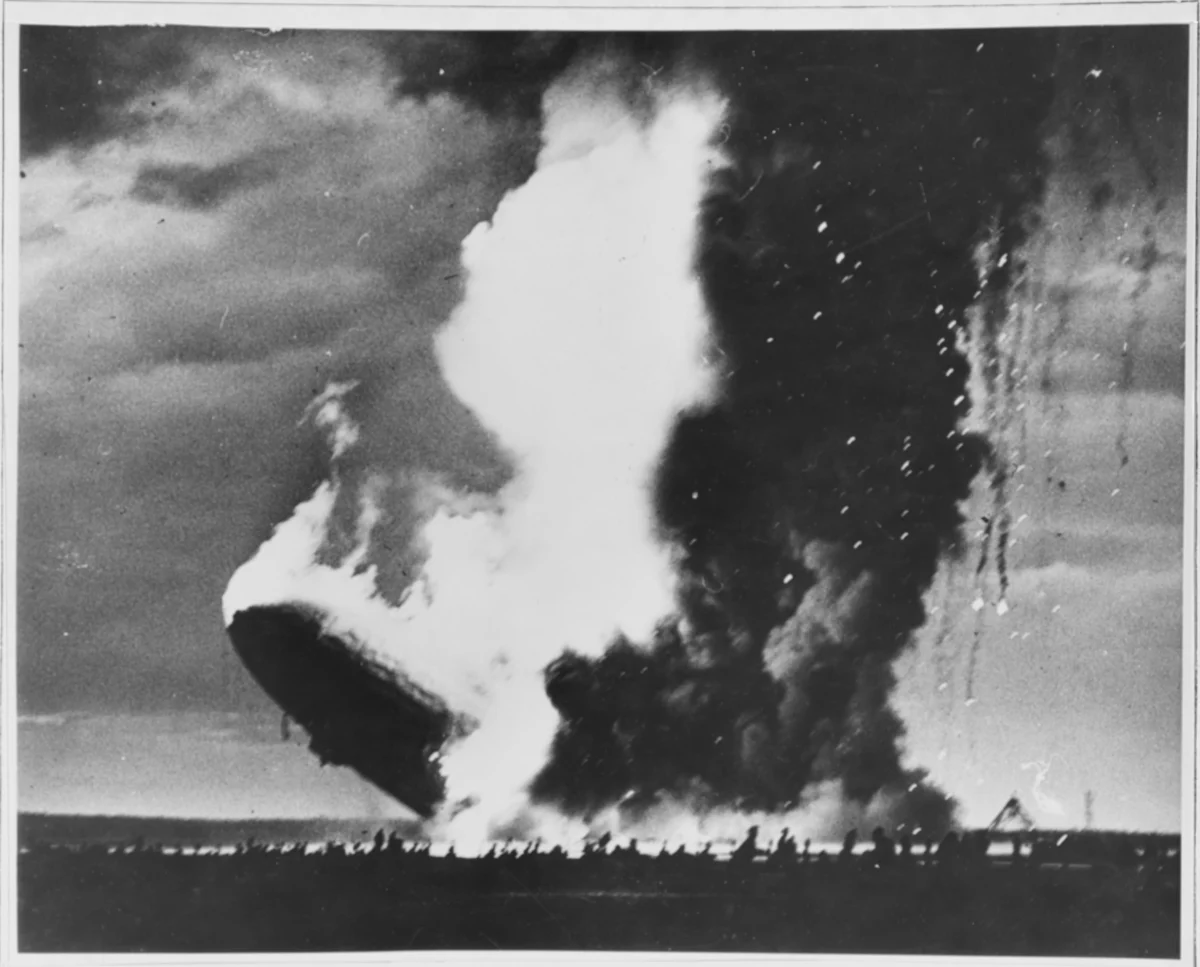 A rigid airship sinking stern first in a huge pale column of flame and dark smoke above a flat field.