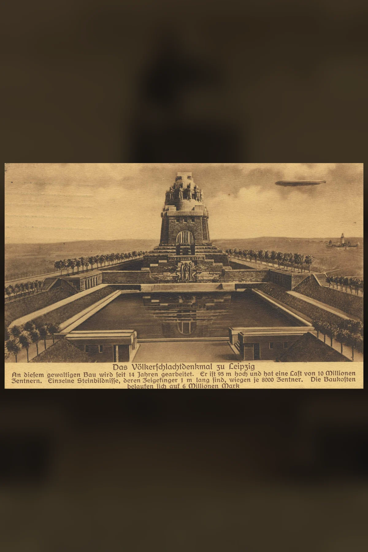 A sepia postcard of a huge stepped stone monument behind a long rectangular pool, with a small rigid airship in the sky at the upper right and two lines of German print along the bottom edge.