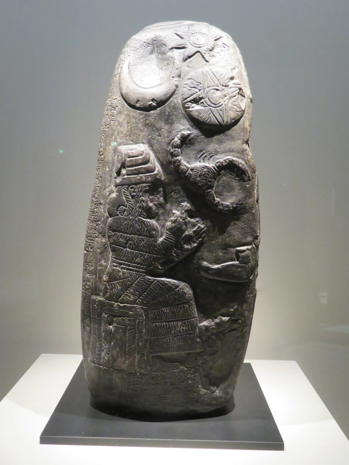 A tall dark stone carved at its top with a crescent, a disc and an eight-pointed star side by side, above figures and writing.