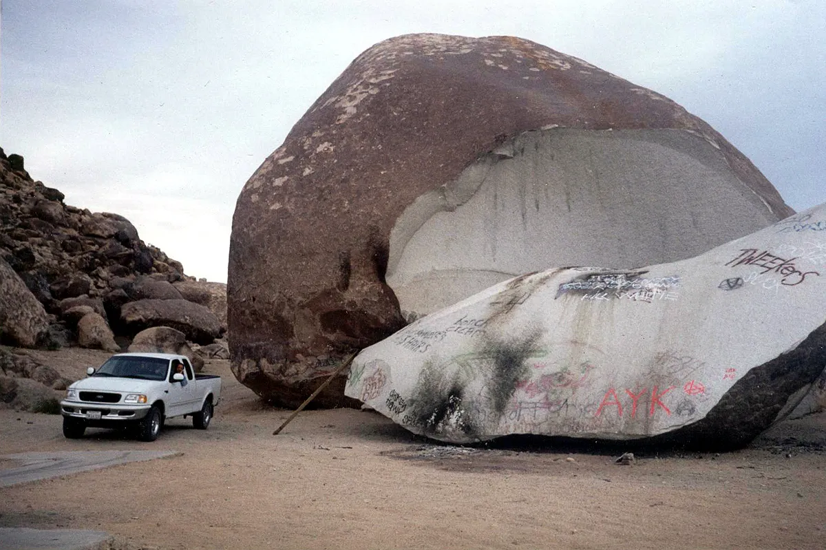 A huge rounded boulder split open, its pale inner face exposed and a graffiti-covered slab lying in front, with a white pickup truck parked beside it for scale.