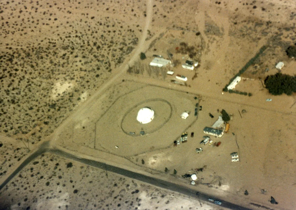 An aerial view of desert scrub with a small white dome inside two oval fence rings, a few low buildings and parked cars beside it, and a paved road crossing the lower part of the frame.