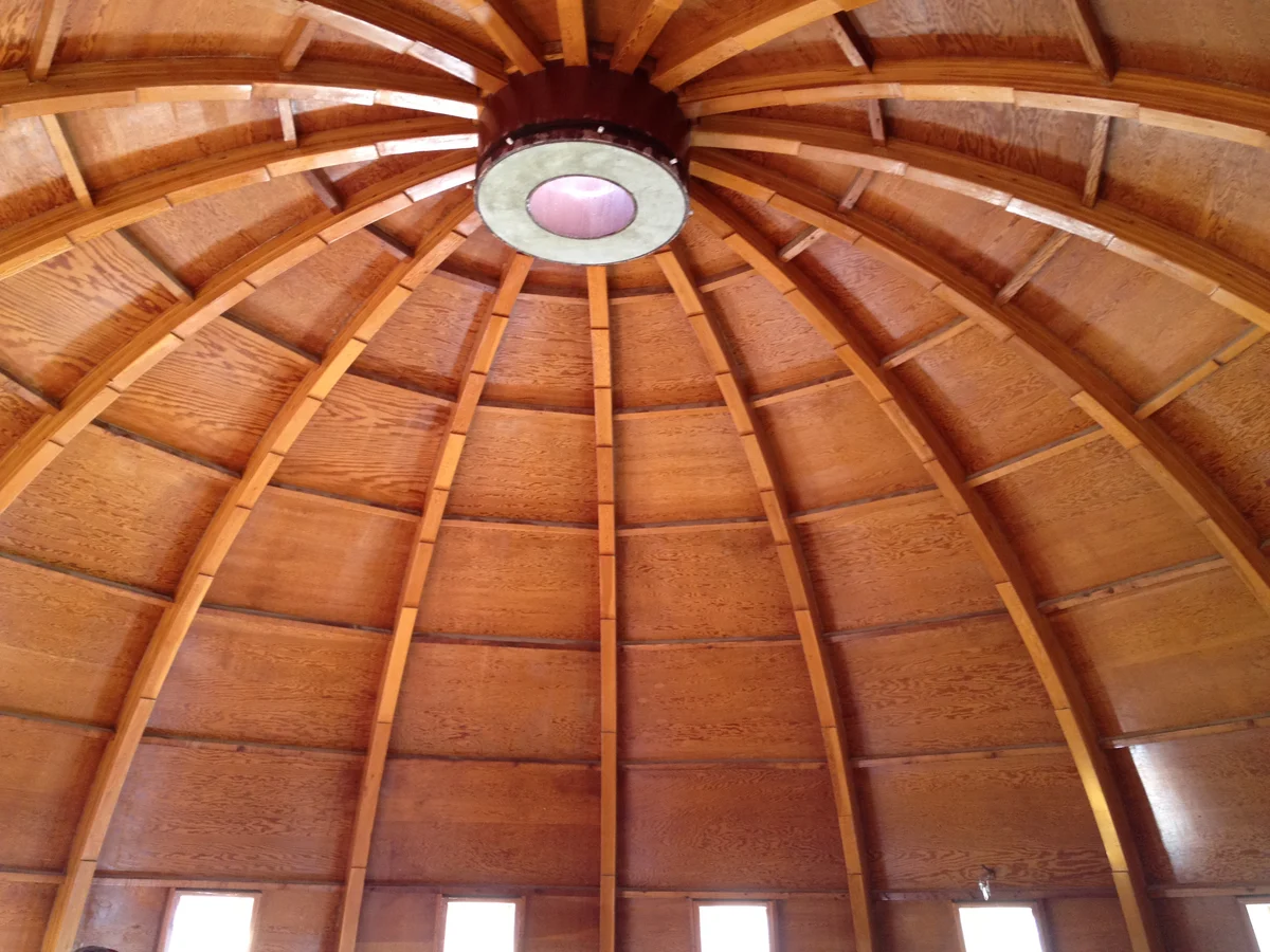 Looking up inside a wooden dome: curved laminated ribs converge on a round concrete ring with a pale disc at its centre, with wood panelling between the ribs and small windows at the base.