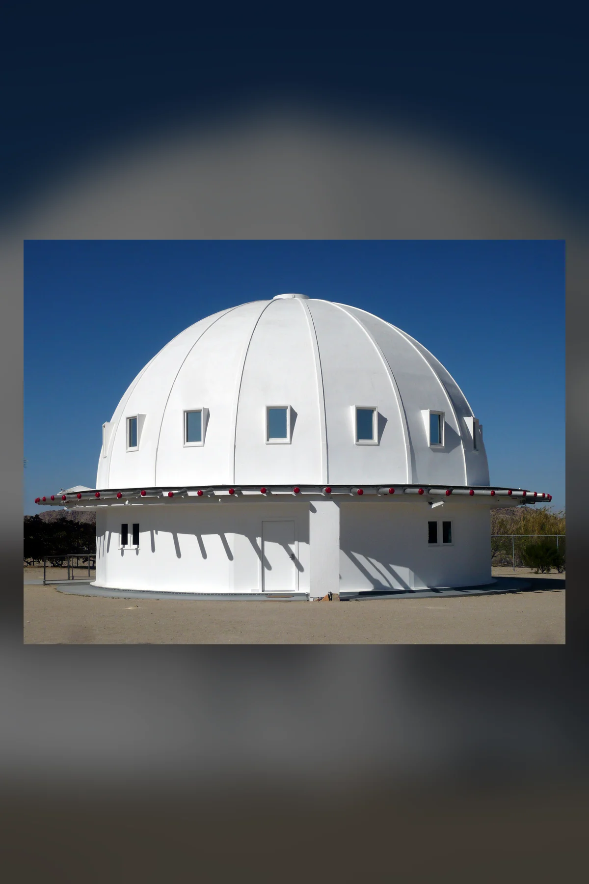 A white dome with sixteen ribs and small square windows rises over a round white base, with a ring of red-tipped rods around the eave, under a clear deep blue sky.