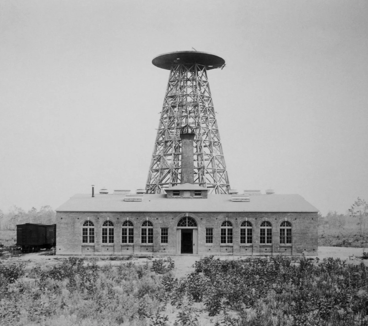The lattice tower with a flat round platform on top, rising behind the brick laboratory, with a coal wagon beside the building and low brush in front.
