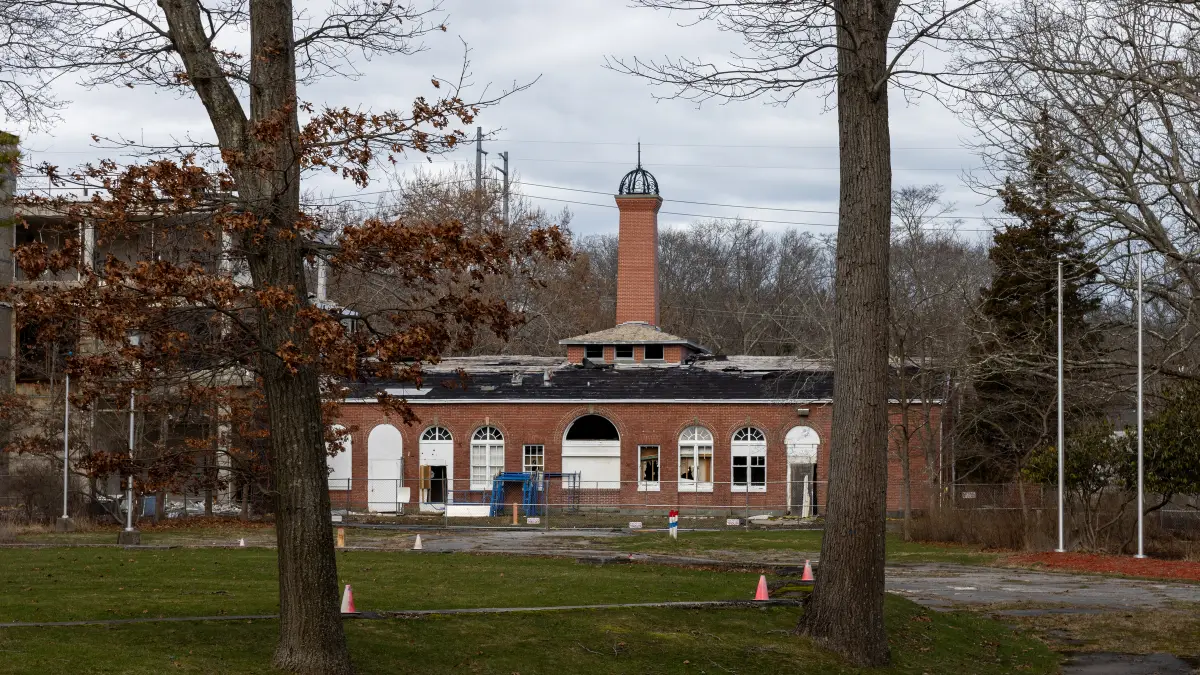 A long red-brick building with arched windows and a tall brick chimney topped by a small cupola, seen across a lawn between bare trees.