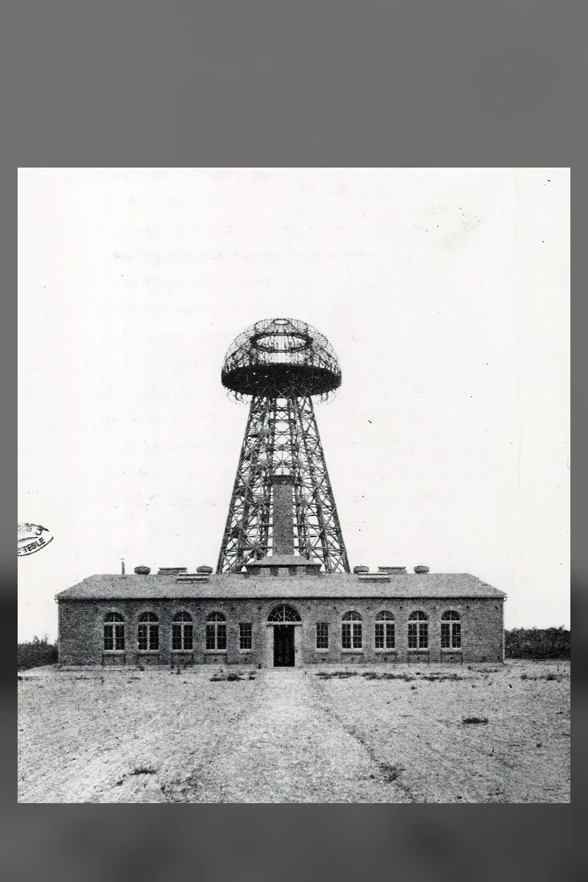 An old black and white photograph of a tall open lattice tower with a round domed top, rising behind a long single-storey brick building with arched windows.