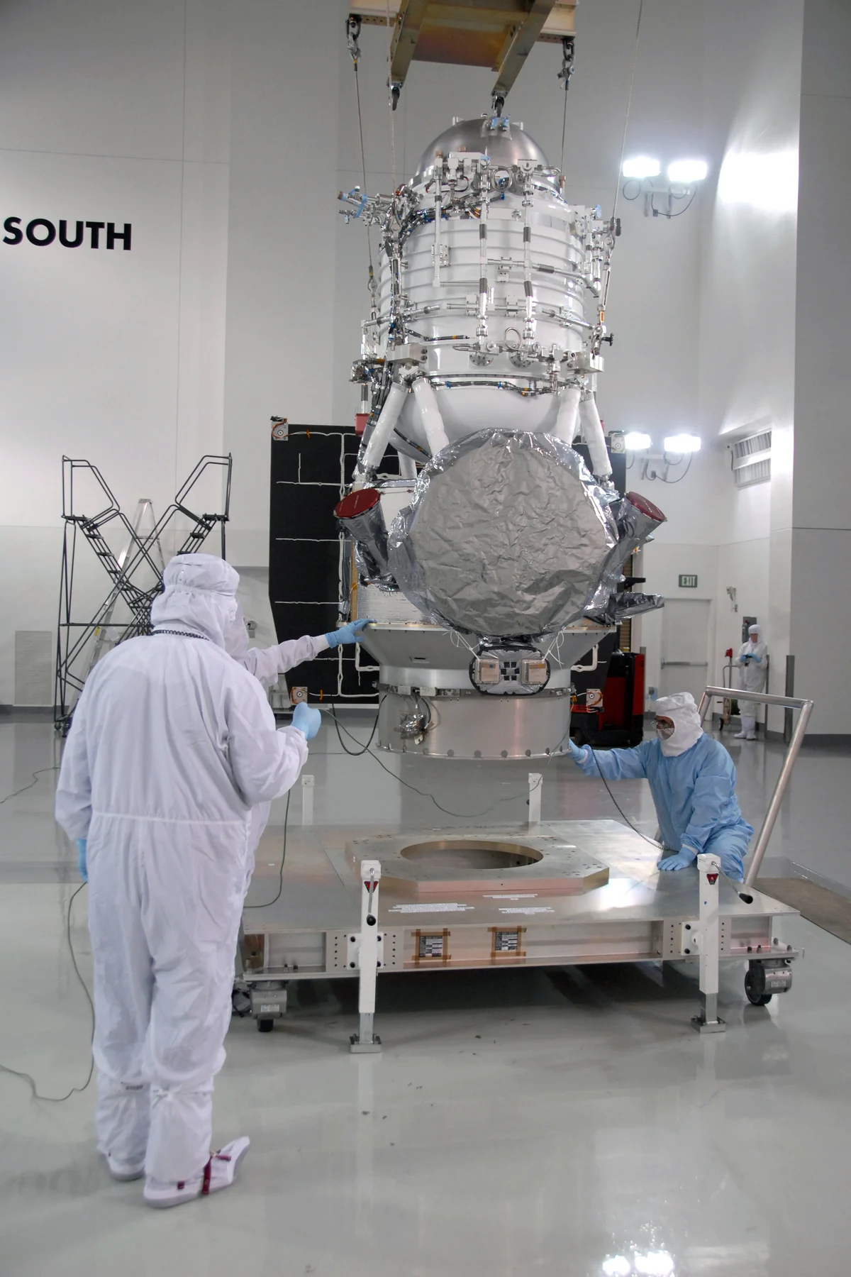 The WISE spacecraft hanging from a crane in a white clean room, its silver cylindrical body above a round grey cover, with technicians in clean-room suits guiding it