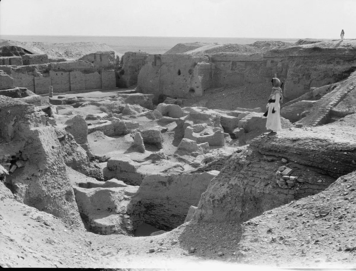 Black and white photograph of deep excavations in mud-brick ruins, a man standing on a high wall for scale.