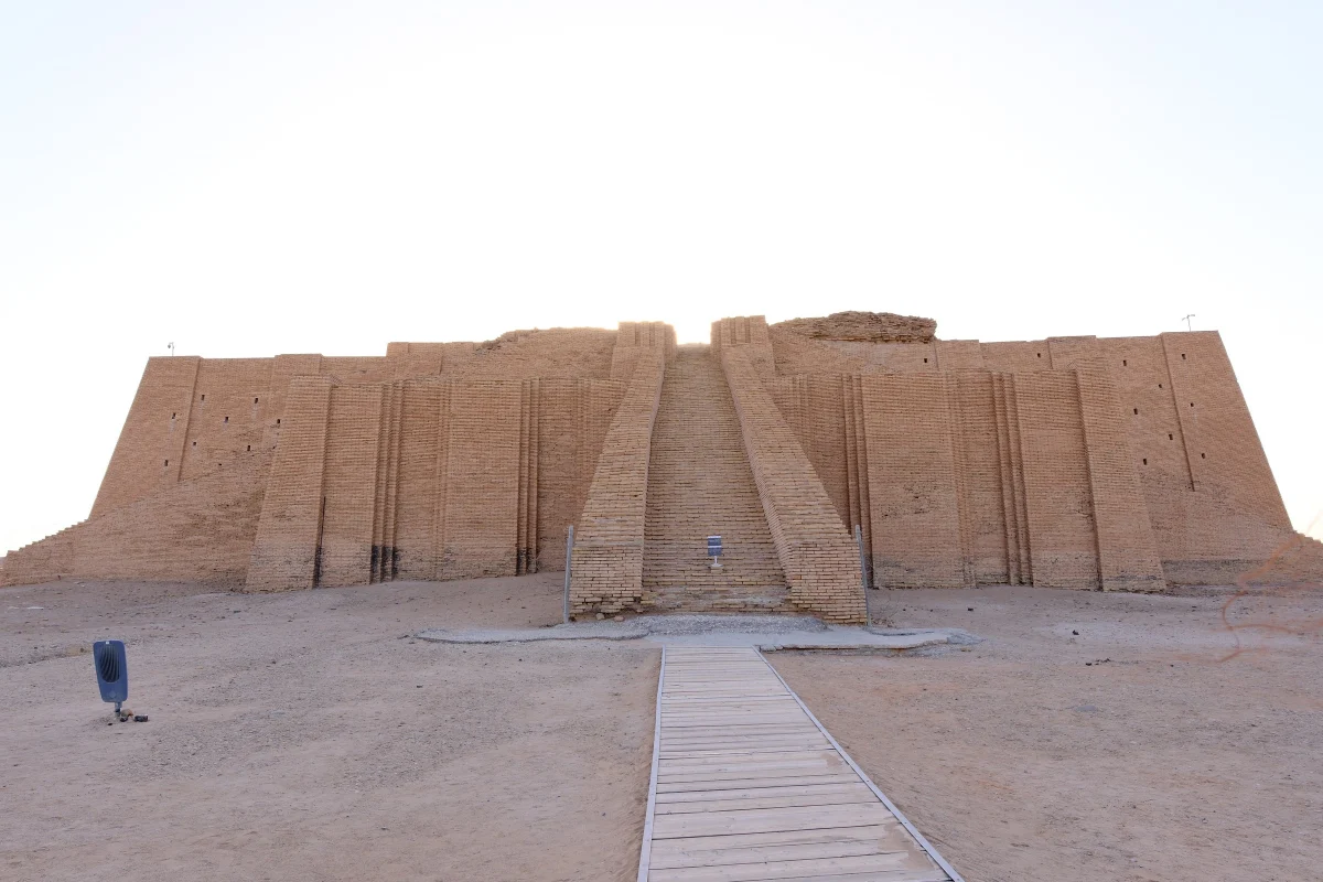 A wide stepped brick temple tower with a central stairway, restored, on a flat sandy plain, a wooden walkway leading to it.