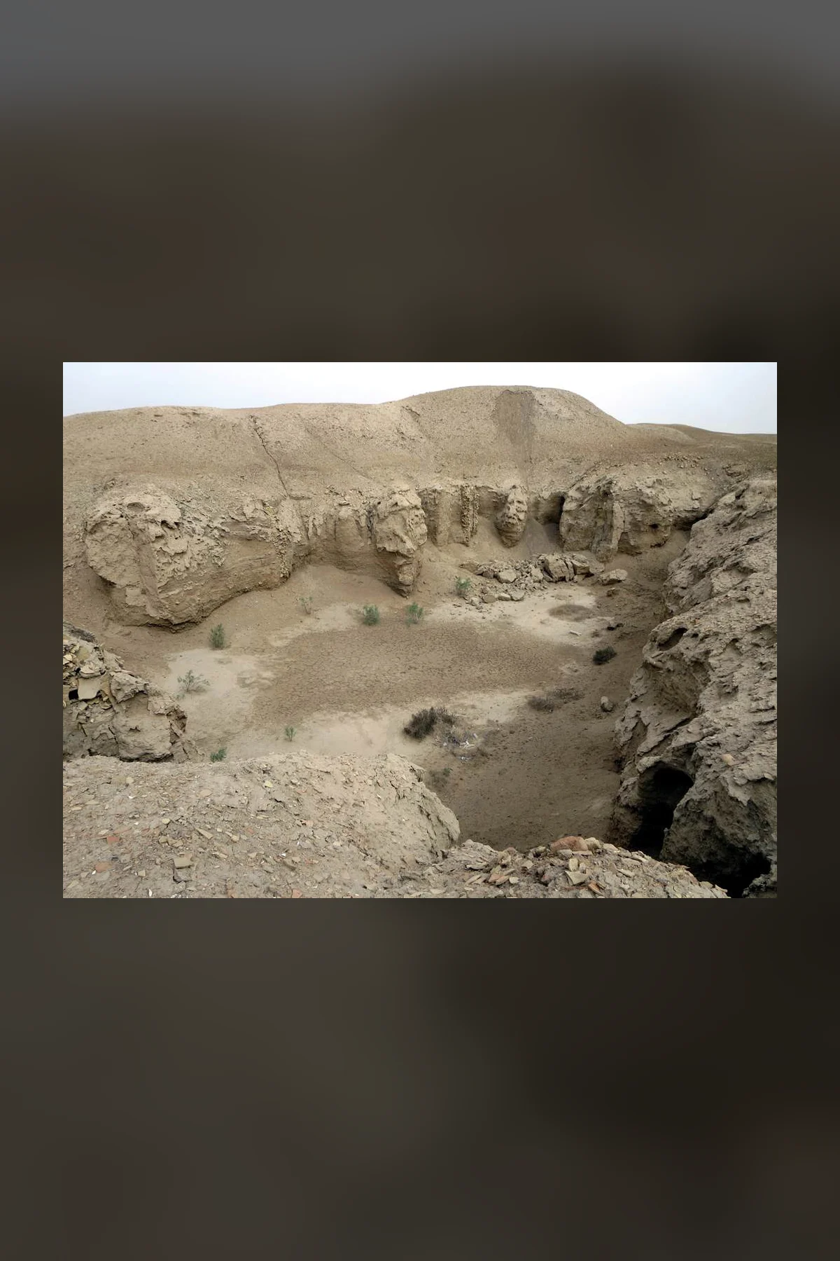 A deep, wide excavation pit in pale desert earth at Ur, its eroded walls of packed earth stepping down to a sandy floor with scattered shrubs.
