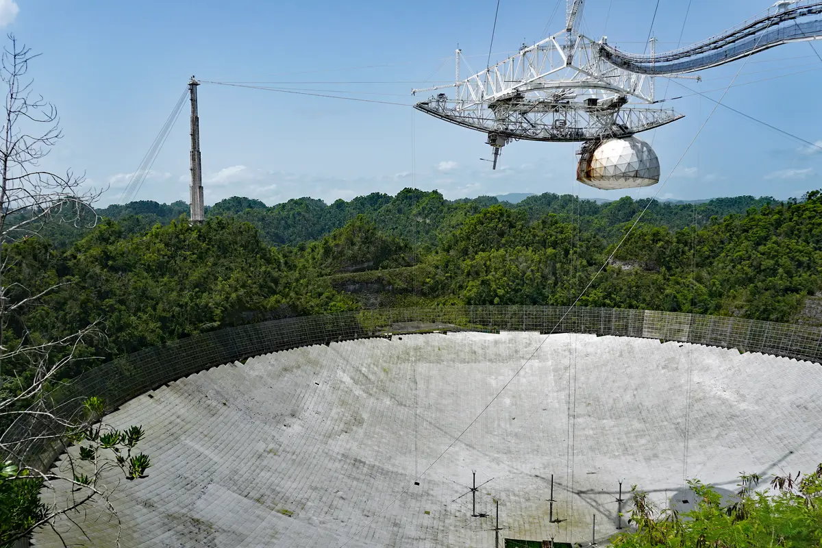 A huge curved dish of mesh panels set into a green tropical hollow, with a triangular instrument platform hanging above it on cables from tall towers.