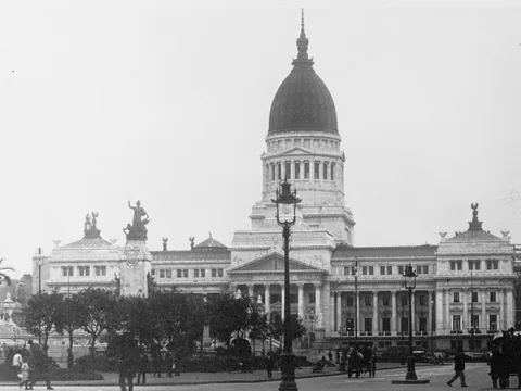 A domed neoclassical parliament with a tall drum, columned portico and flanking wings, street lamps and a statue group in front, people on the pavement.
