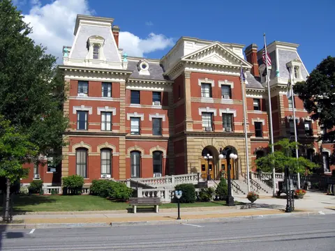 Colour photo from the street: a three-storey red brick courthouse with stone trim, mansard-roofed corner pavilions, a central arched porch and flagpoles.
