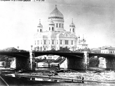 Black and white photograph of a white cathedral with a large central dome and four smaller domes, seen beyond the arches of a river bridge.