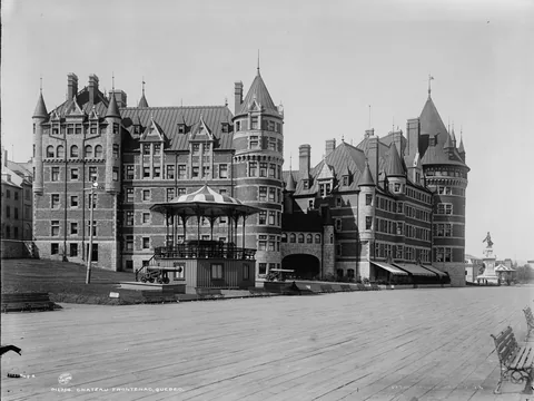 A turreted brick chateau hotel with steep roofs, a domed bandstand and cannon on a wide boardwalk terrace in front of it.