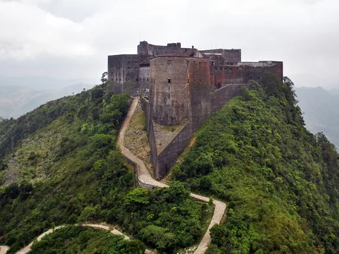 A dark stone mountain fortress with a prow-like bastion on a green ridge, a winding path climbing to it, mist on the peaks behind.