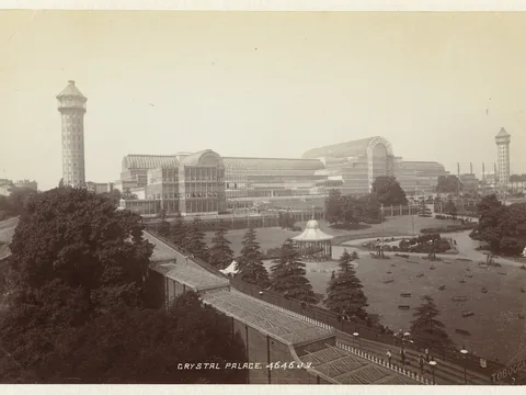 A vast glass and iron exhibition palace with barrel-vaulted roofs and two tall water towers, seen across terraced gardens with a bandstand and paths.