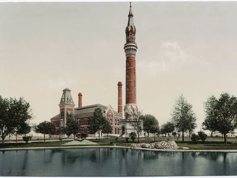 Colour print of a tall slim red brick tower with a stone base and spire, beside a brick pump house with chimneys, trees and a pond in front.
