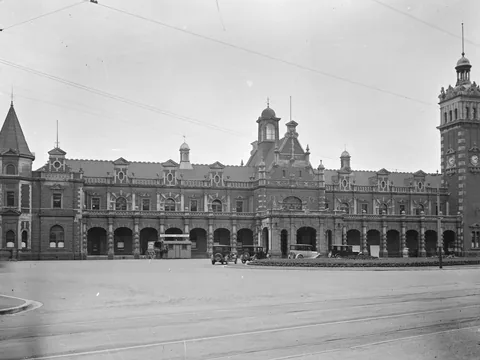 Black and white photograph of a long brick and stone station with arcaded front, a tall clock tower at right and a pointed turret at left, with 1920s cars outside.