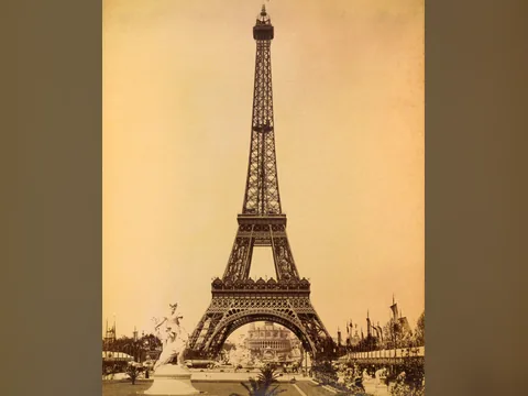 The complete iron lattice tower seen straight on along its axis, an exhibition palace visible through the great arch, a white statue and flags in the foreground.