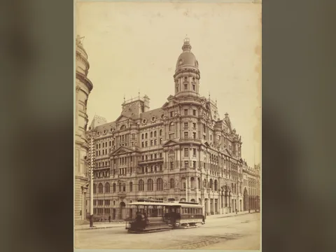 Sepia photograph of a huge ornate corner hotel of seven floors under a domed tower, with a horse tram passing on the street in front.