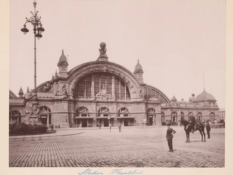 Sepia photograph of a stone station front under a great arched window, with a globe statue on top, a tall lamp standard at left, and a man and a horseman in the square.