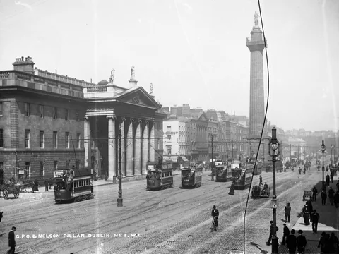 A wide stone post office with a columned portico and roof statues on a busy street, electric trams passing and a tall column rising beyond it.