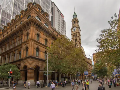 Colour photograph of a long sandstone building with a clock tower rising beyond trees, along a pedestrian street busy with people.