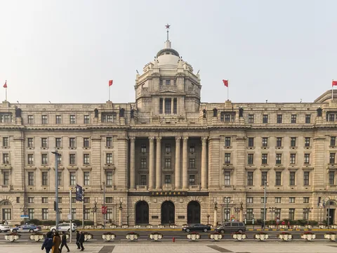 A broad granite bank facade with a central columned portico and a dome, seen straight on across the Bund road, red flags along the roof.