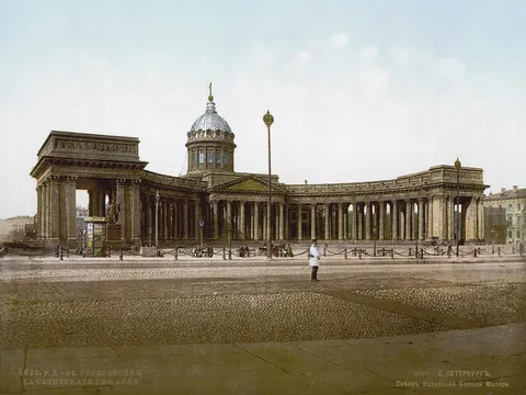 Colour photochrom of a domed cathedral behind a long curved colonnade, seen across an open cobbled square, with a lone policeman in white in the foreground.